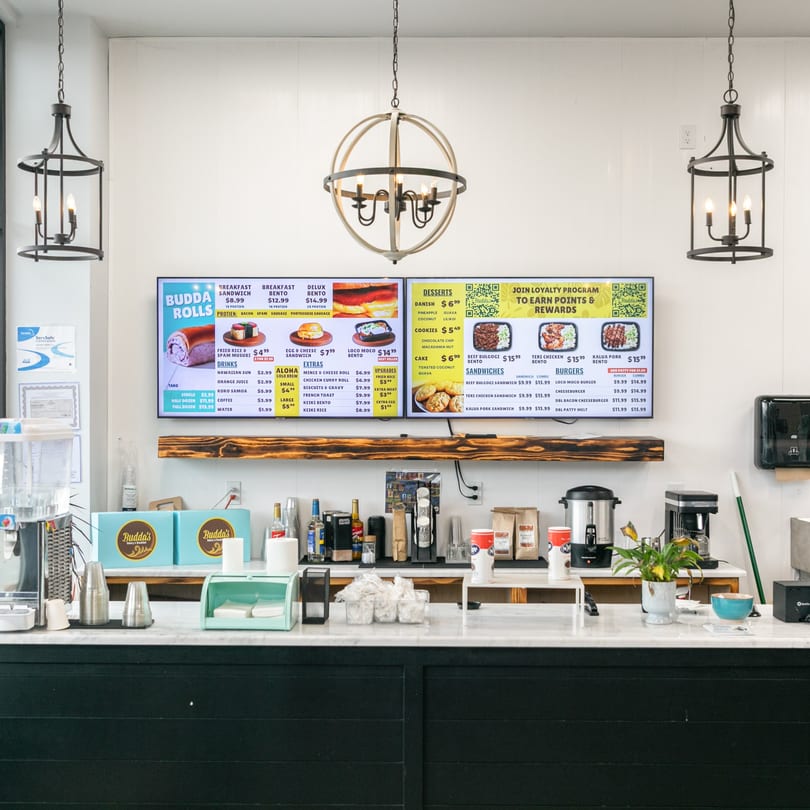 A clean counter at Budda's Hawaiian Bakery & Grill with digital menu boards displaying Hawaiian dishes.