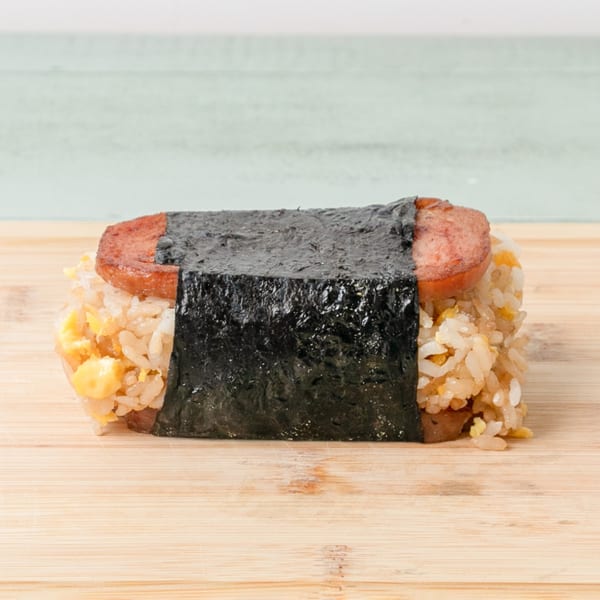 A Budda's Hawaiian Bakery & Grill Spam Musubi with rice and egg, wrapped in nori, on a wooden board.