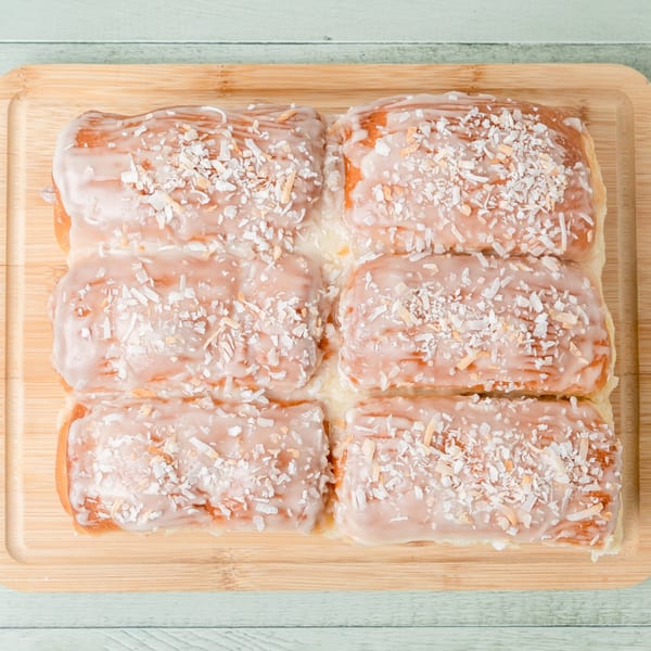 Six Budda's Hawaiian Bakery & Grill coconut rolls with white icing and shredded coconut on a wooden board.