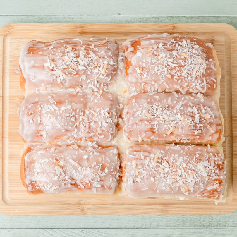Six Budda's Hawaiian Bakery & Grill coconut rolls with white icing and shredded coconut on a wooden board.
