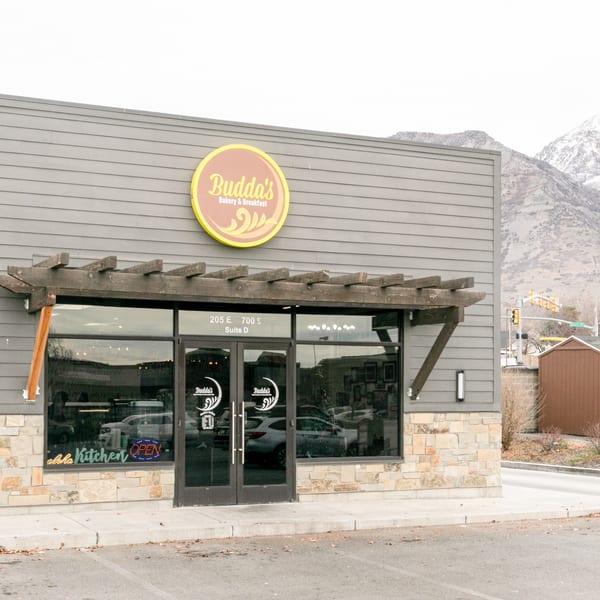 Exterior of Budda's Hawaiian Bakery & Grill with a yellow sign and snow-capped mountains in the background.