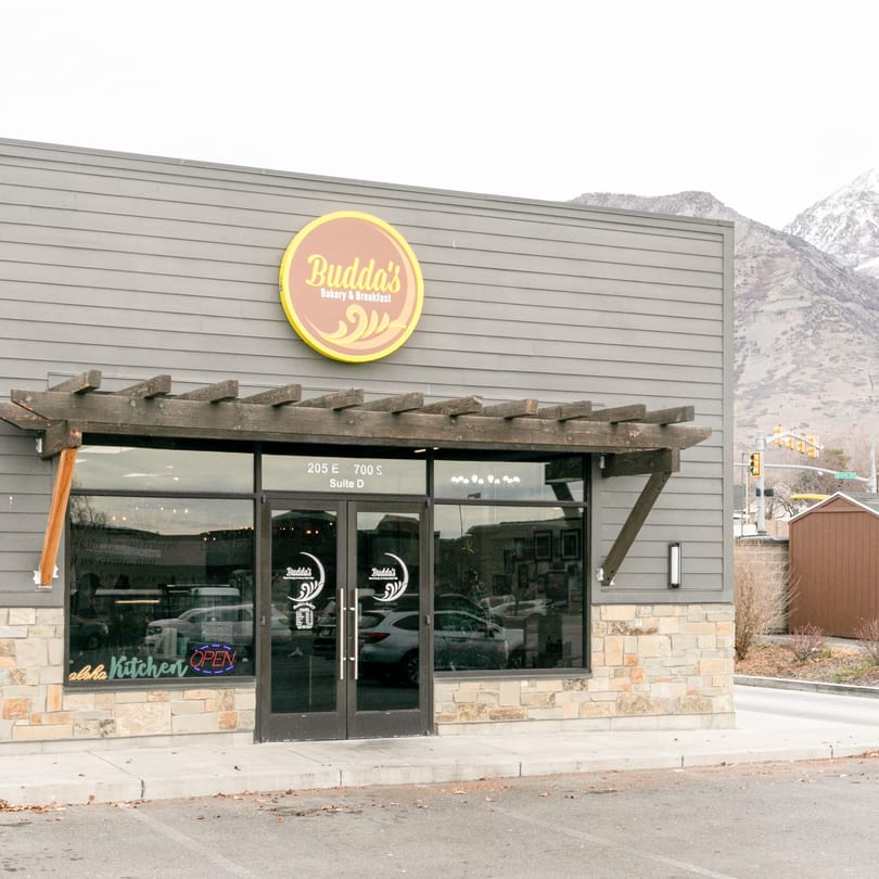 Exterior of Budda's Hawaiian Bakery & Grill with a yellow sign and snow-capped mountains in the background.