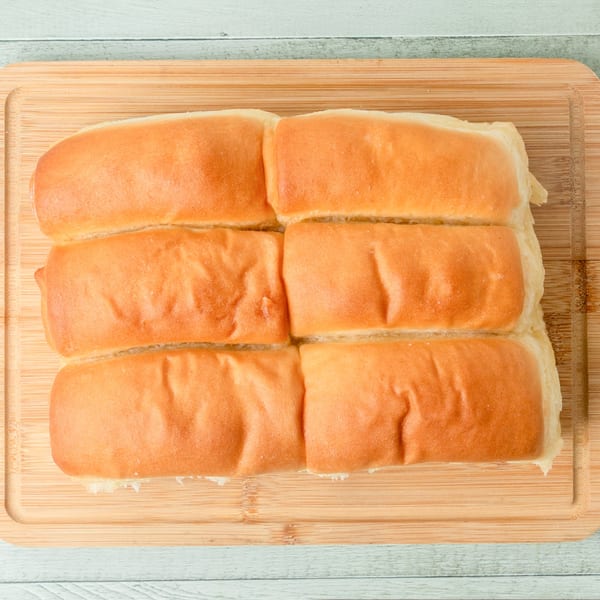 Six golden-brown Hawaiian sweet rolls on a light wooden cutting board.